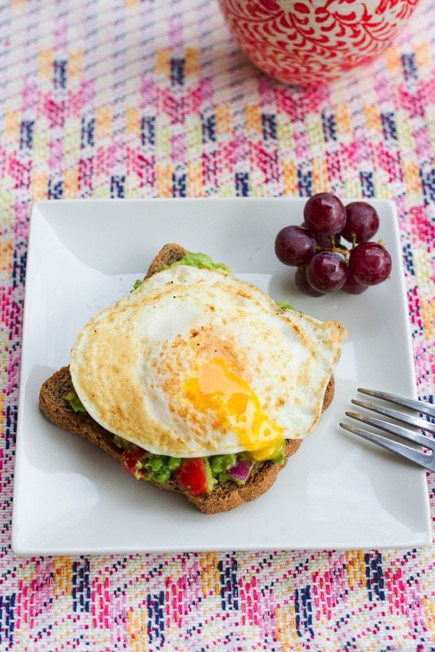 A fried egg resting on top of a slice of toast with spread with guacamole. The yolk has been poked and is drizzling out of the egg.