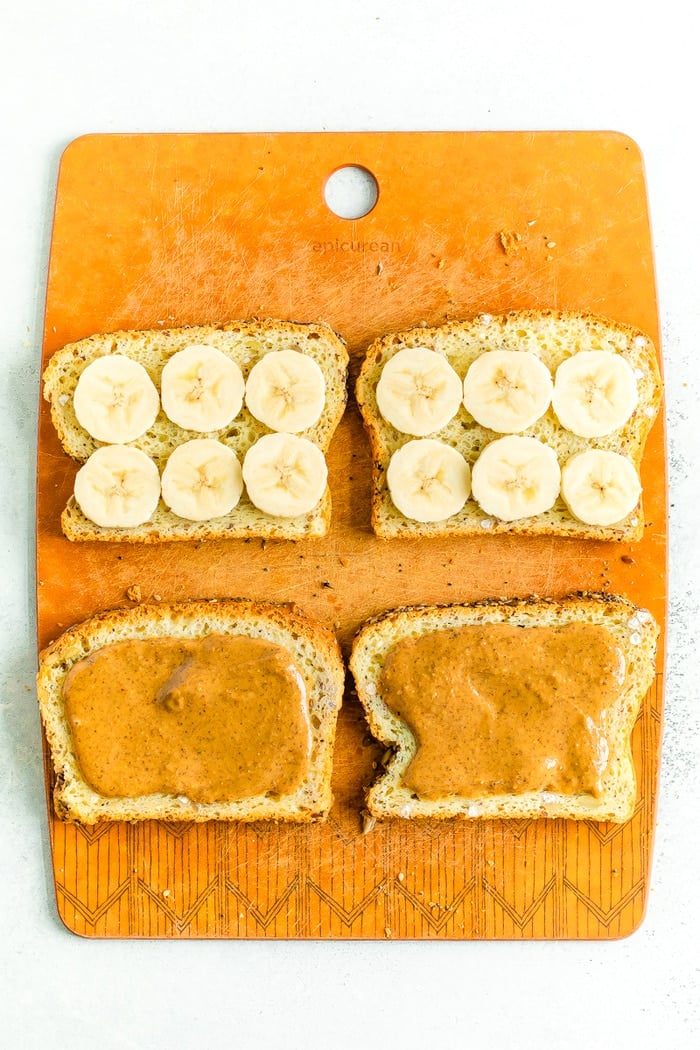 Cutting board with two slices of bread with banana slices, and two slices with almond butter.