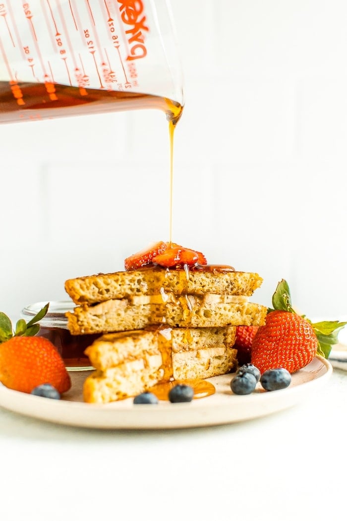 Pyrex measuring cup pouring maple syrup over two slices of stuffed french toast on a plate with berries.