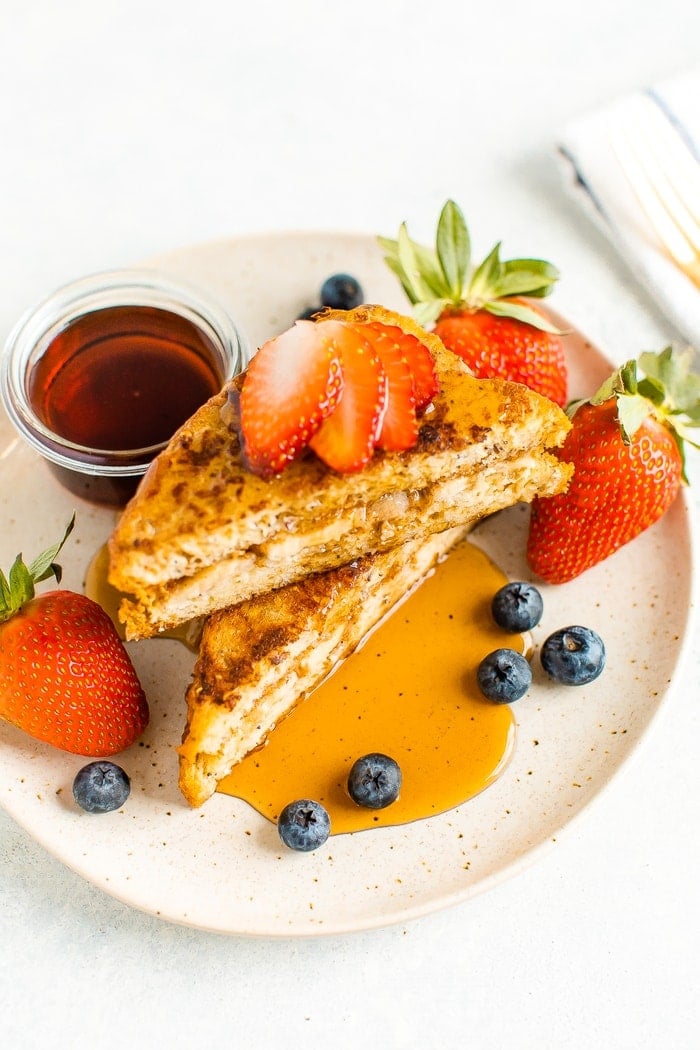 Banana and almond butter stuffed french toast on a plate with berries and topped with maple syrup.