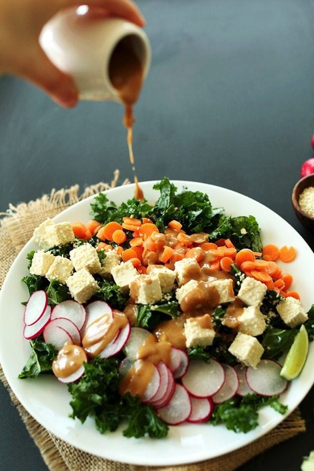 Crunchy Thai Kale Salad on a white plate. Hand in upper left corner pouring dressing over salad