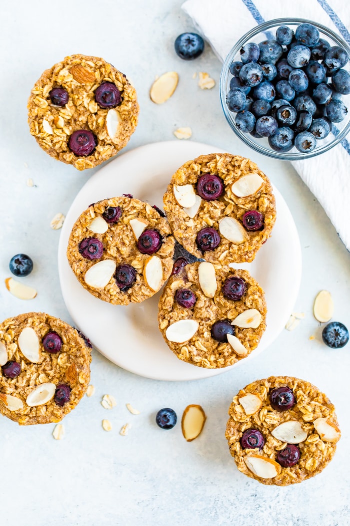 Plate with blueberry baked oatmeal cups and oatmeal cups, blueberries, a bowl of blueberries and almond slivers are around on the table.