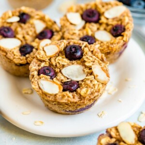 Plate with blueberry baked oatmeal cups and oatmeal cups, blueberries, a bowl of blueberries and almond slivers are around on the table.