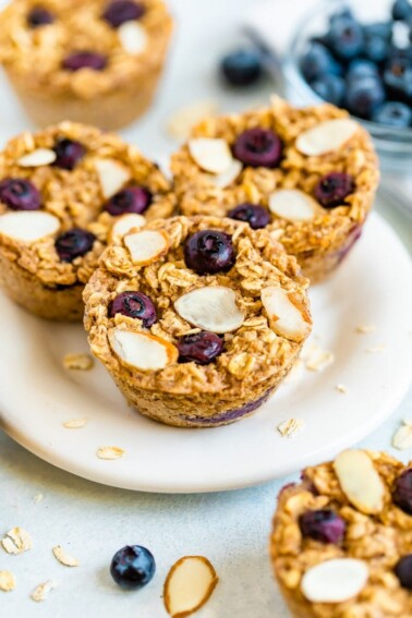 Plate with blueberry baked oatmeal cups and oatmeal cups, blueberries, a bowl of blueberries and almond slivers are around on the table.