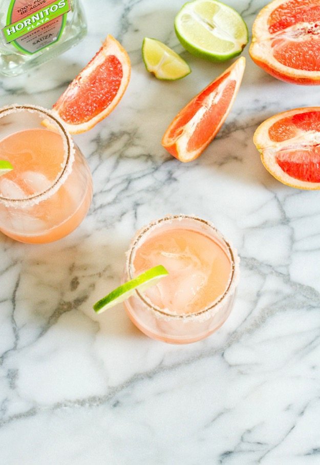 Paloma cocktails in glasses with lime wedges. A bottle of tequila and slices of grapefruit are on the counter.