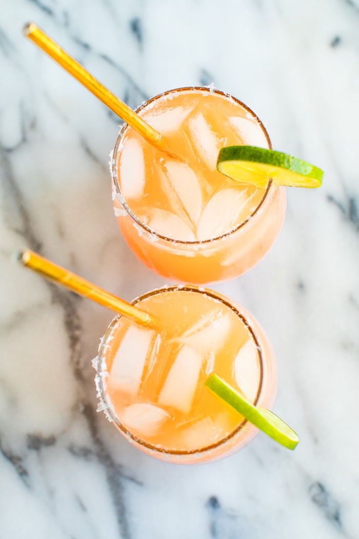 Two paloma cocktails in stemless wine glasses with ice, lime wedges and gold straws.