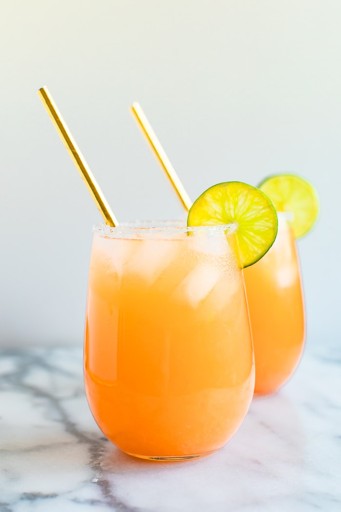 Two paloma cocktails in stemless wine glasses with ice, lime wedges and gold straws.