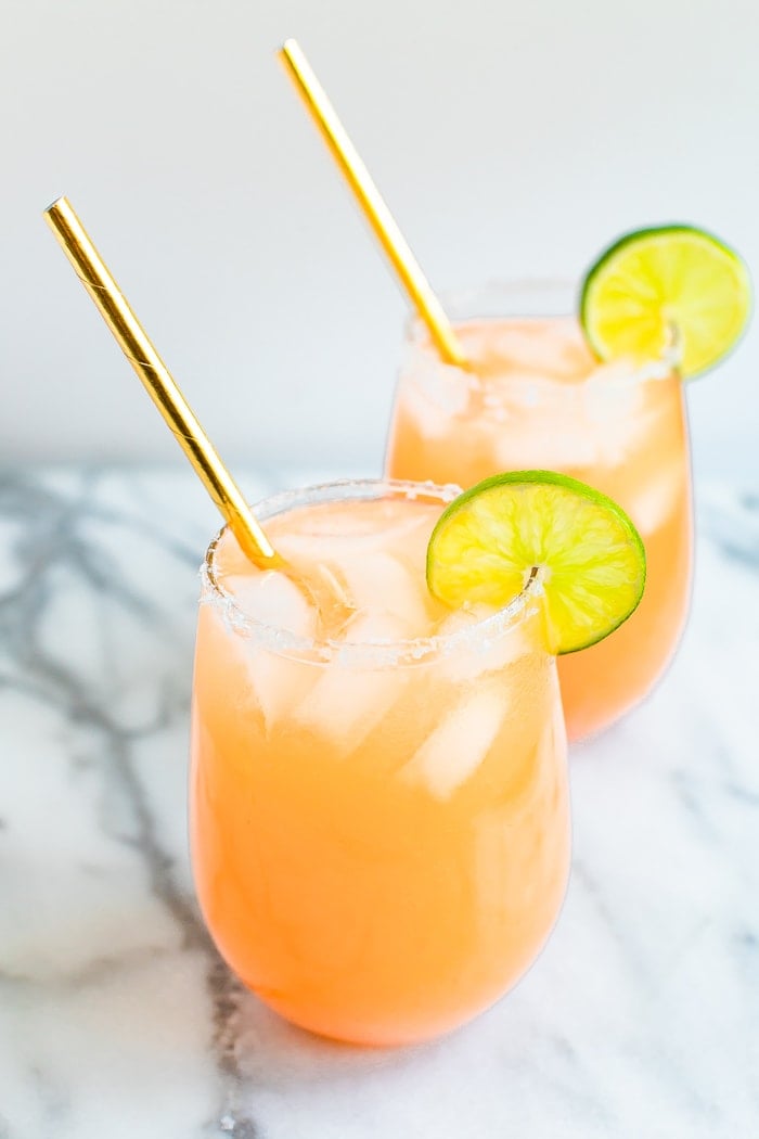 Two paloma cocktails in stemless wine glasses with ice, lime wedges and gold straws.