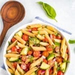 Bowl of clean eating penne pasta salad mixed with basil, cherry tomatoes, cucumber and red onion. Next to the bowl are fresh basil leaves and a wooden mixing spoon.