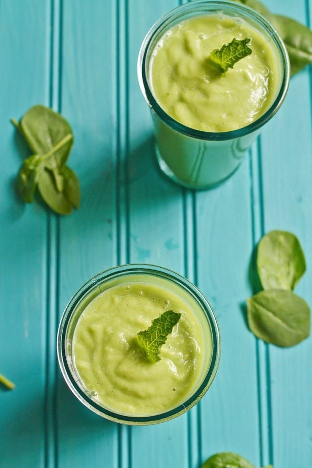 Two pear smoothies in glasses on a teal background with spinach leaves around the glasses. 