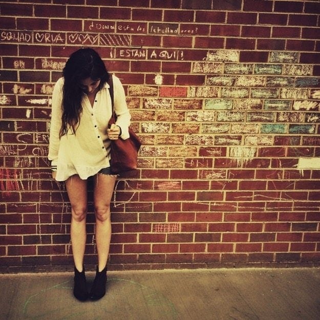 Girl with long brown hair in white top and jean shorts standing against a red brick wall. 