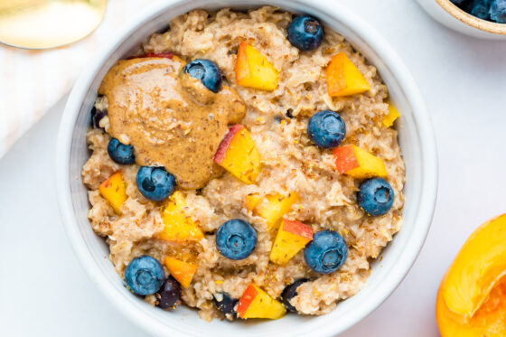 Blueberry peach oatmeal in bowl with blueberries and fresh chopped peaches on top with a little almond butter on top as well.