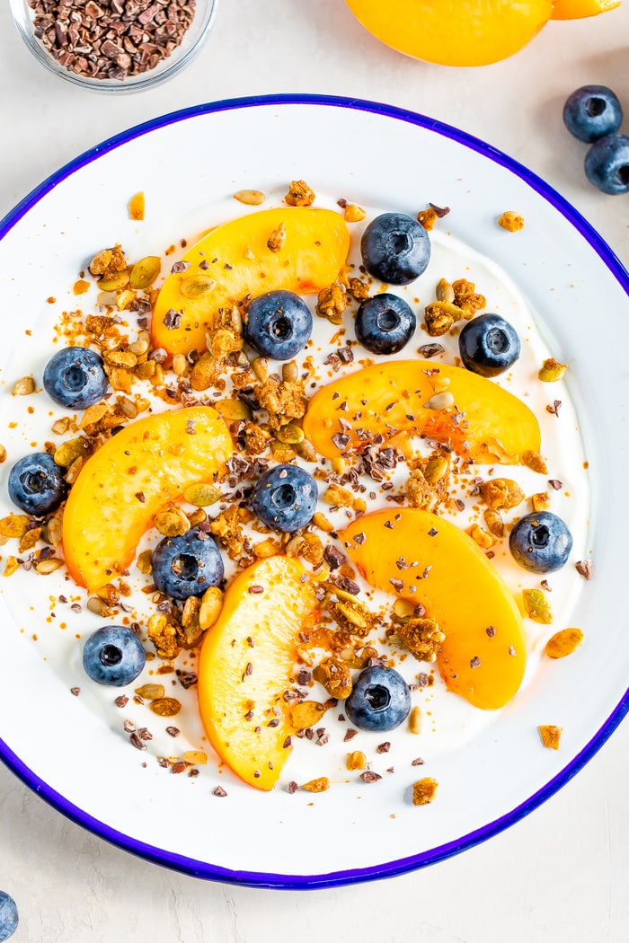 Birds eye view of a bowl of yogurt with fresh peach slices and blueberries, topped with granola, cacao nibs and nut butter.