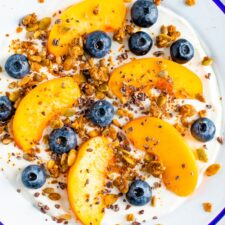 Bowl of yogurt with fresh peach slices and blueberries, topped with granola, cacao nibs and nut butter.