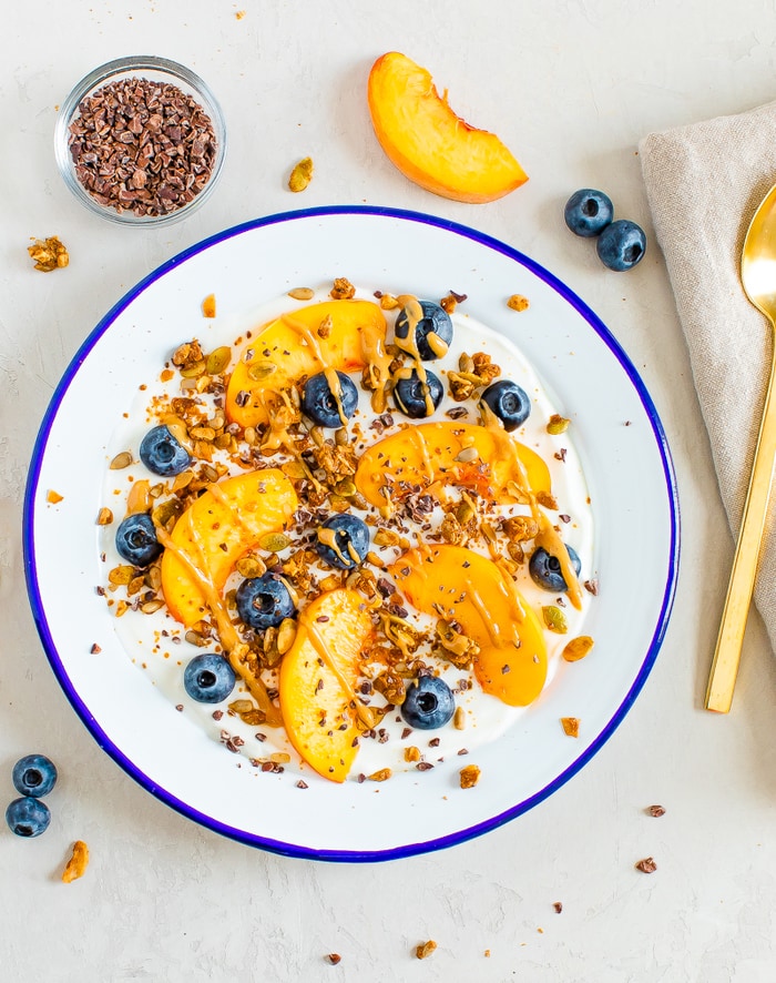 Birds eye view of Bowl of yogurt with fresh peach slices and blueberries, topped with granola, cacao nibs and nut butter. Bowl of cacao nibs, a slice of peach and a few blueberries are around the bowl.