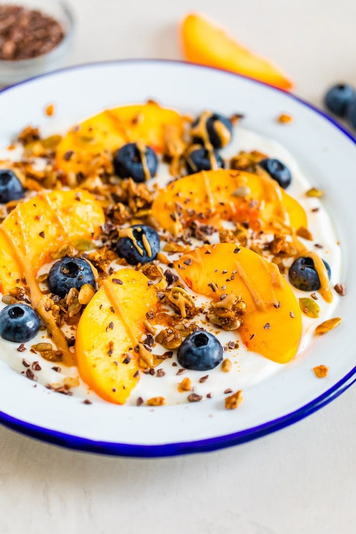 Bowl of yogurt with fresh peach slices and blueberries, topped with granola, cacao nibs and nut butter.