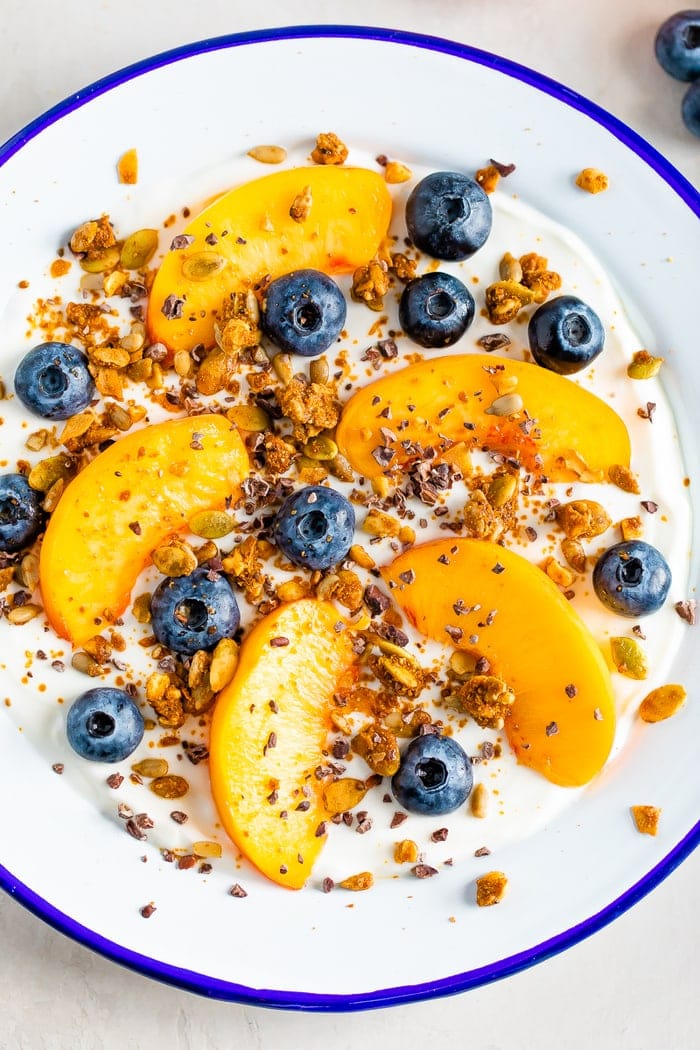 Bowl of yogurt with fresh peach slices and blueberries, topped with granola, cacao nibs and nut butter.