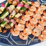 Grilled skewers of shrimp and zucchini, onion, and mushrooms on a navy serving tray.