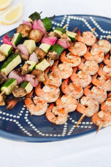 Grilled skewers of shrimp and zucchini, onion, and mushrooms on a navy serving tray.