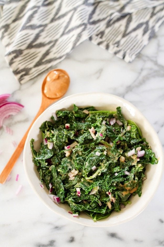 Kale Salad with Peanut Dressing -- Tender kale with a rich and spicy peanut dressing and chopped red onion! This salad is packed with umami flavor and a fabulous side dish for almost any meal. #vegan #glutenfree An overhead photo of a bowl of kale salad with spicy peanut dressing with a wooden spoon laying next to it.