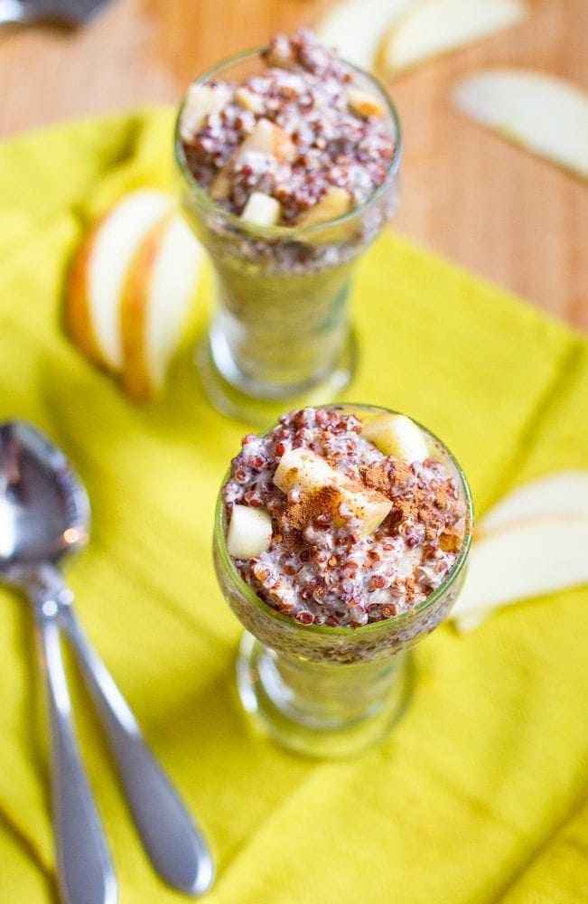 Two tall, narrow glass jars with apple cinnamon overnight quinoa. There are two spoons and some apple slices on the table.