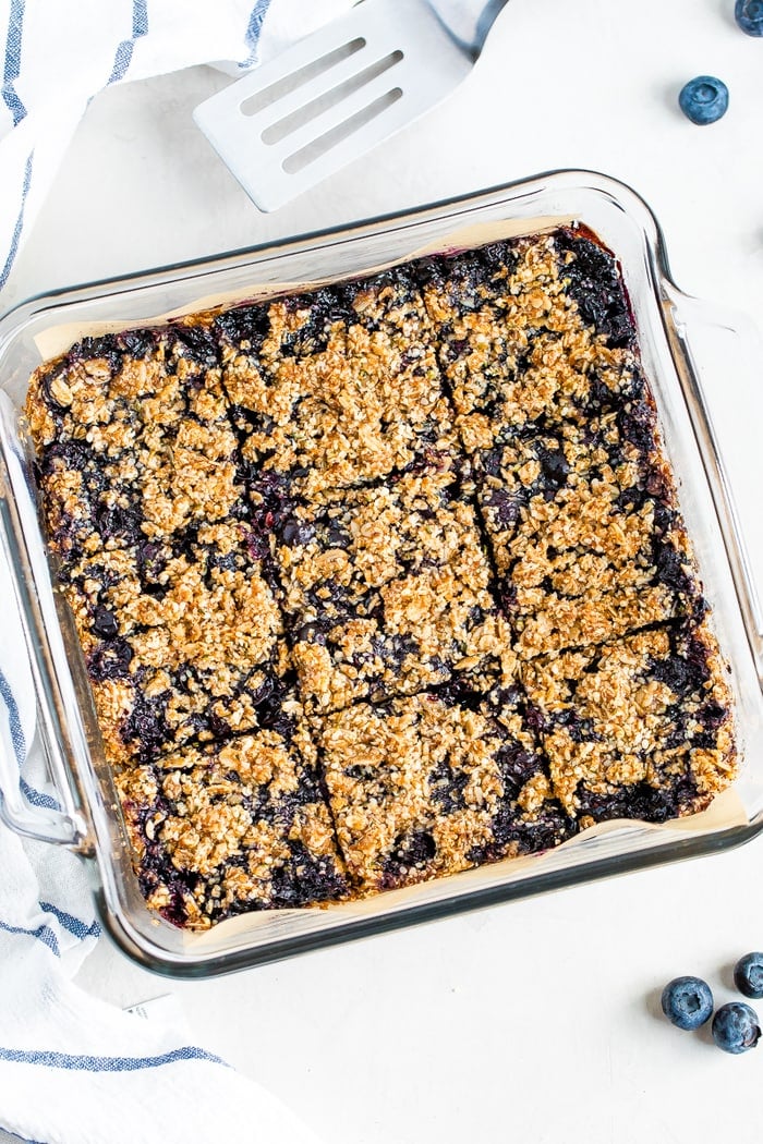 A square glass baking dish with blueberry crumble bars cut into 9 squares. A spatula, dish cloth, and blueberries are around the dish.