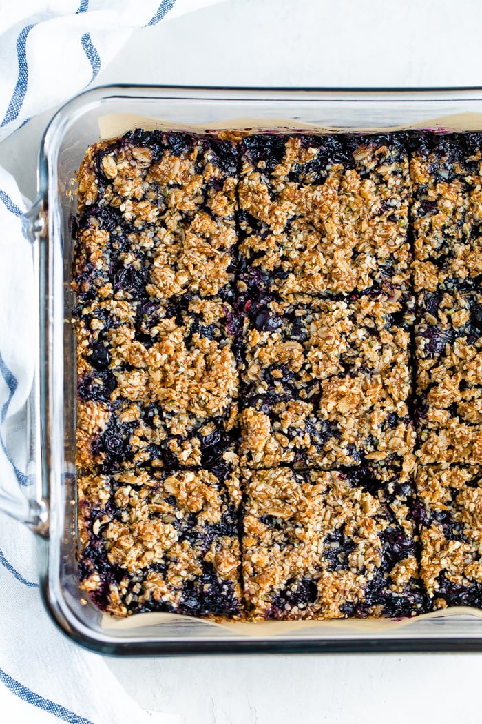 A square glass baking dish with blueberry crumble bars cut into 9 squares. A dish cloth is around the dish.