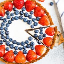 Greek yogurt tart decorated with strawberries and blueberries. A striped dish towel, sliced strawberries, and blueberries surround the tart. A slice is cut for one piece in the tart.