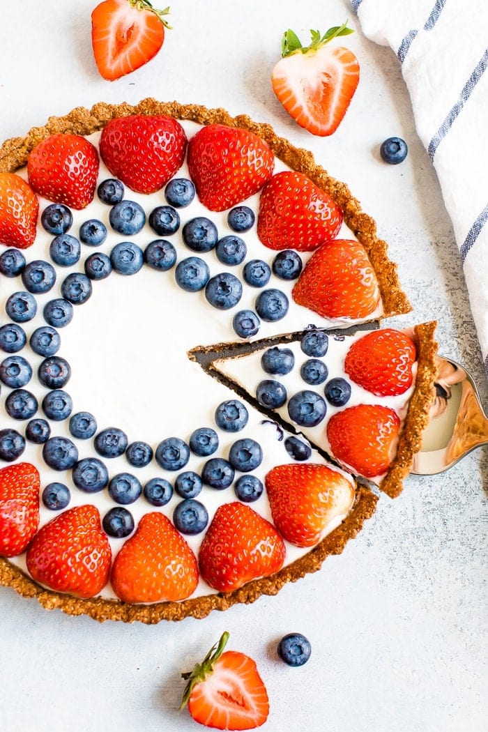 Greek yogurt tart decorated with strawberries and blueberries. A striped dish towel, sliced strawberries, and blueberries surround the tart. A slice is cut for one piece in the tart.