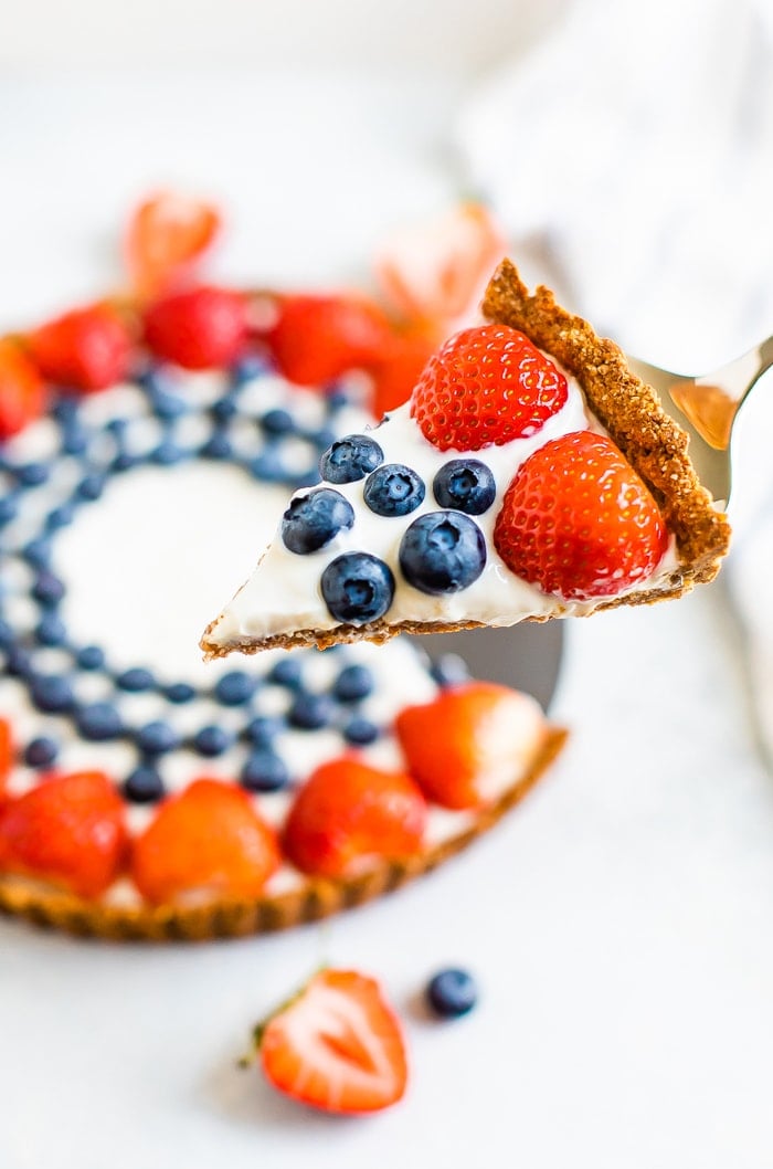 Silver pie spatula holding a slice of greek yogurt berry tart topped with strawberries and blueberries. The rest of the tart is in the background.