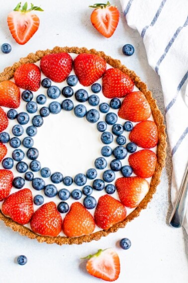 Greek yogurt tart decorated with strawberries and blueberries. A striped dish towel, sliced strawberries, and blueberries surround the tart.