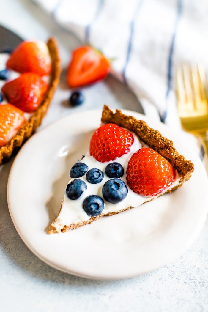 Slice of greek yogurt berry tart with strawberries and blueberries on a plate. A fork, striped napkin, and tart are beside the plate.