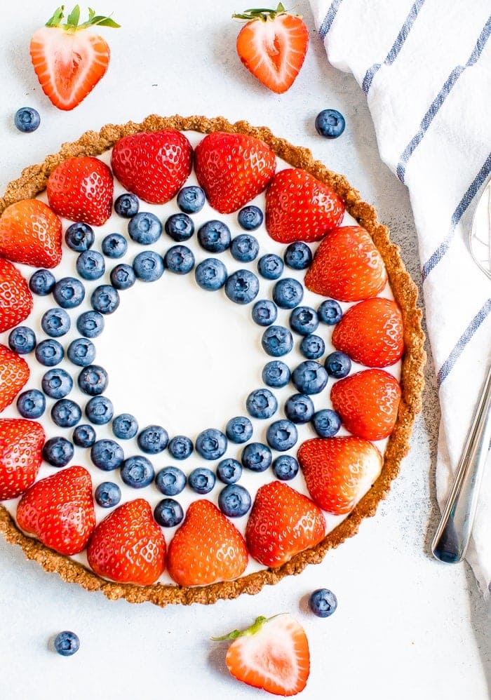 Greek yogurt tart decorated with strawberries and blueberries. A striped dish towel, sliced strawberries, and blueberries surround the tart.