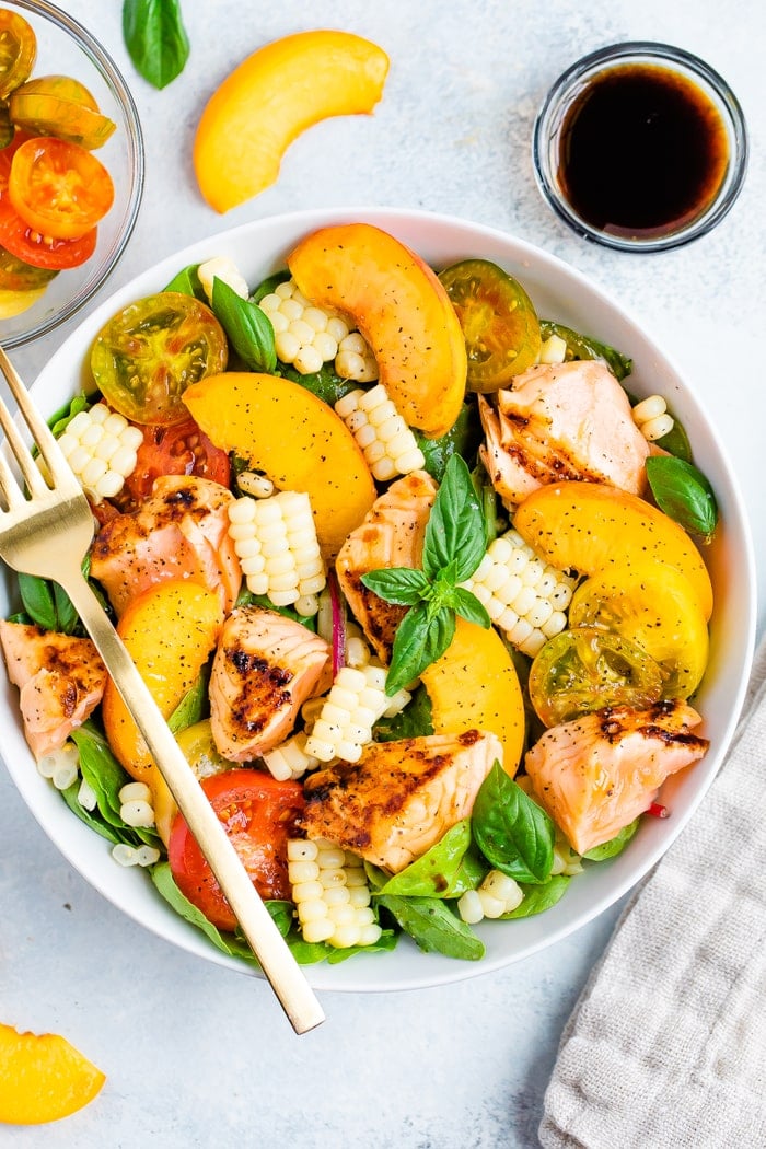 Salad topped with grilled salmon, corn, basil, peaches, and tomatoes. A fork is on the plate and a bowl of tomatoes and a container of dressing are on the side.