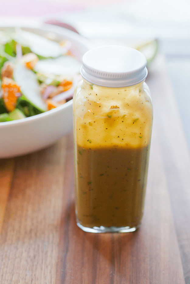 A closed glass container with Avocado Balsamic Dressing. A bowl of salad is in the background.