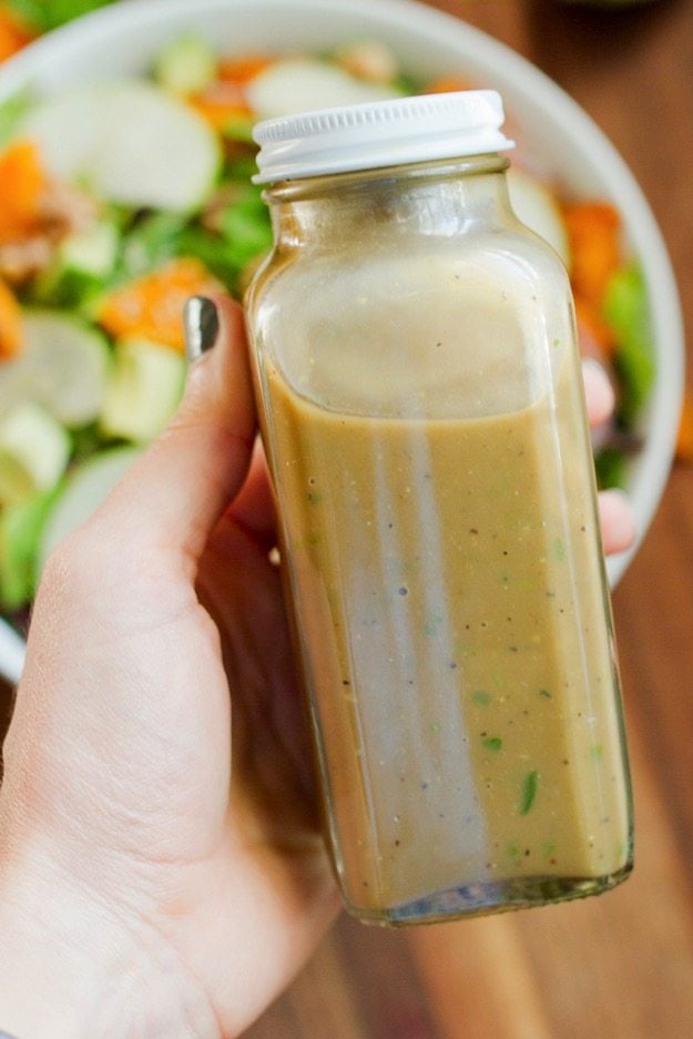 A hand holding up a closed container of Avocado Balsamic Dressing. There is a salad in the background.