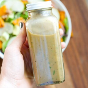 A hand holding up a closed container of Avocado Balsamic Dressing. There is a salad in the background.