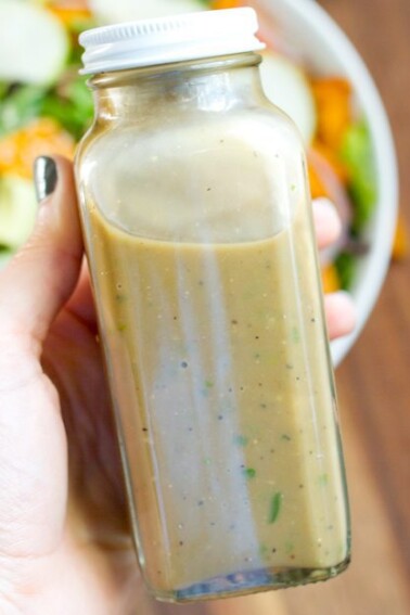 A hand holding up a closed container of Avocado Balsamic Dressing. There is a salad in the background.