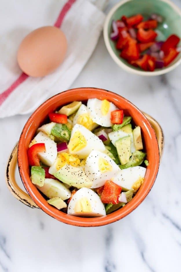 One bowl with chopped hardboiled eggs, avocado, and vegetables, another bowl with chopped peppers and onions, hardboiled egg on the side. 