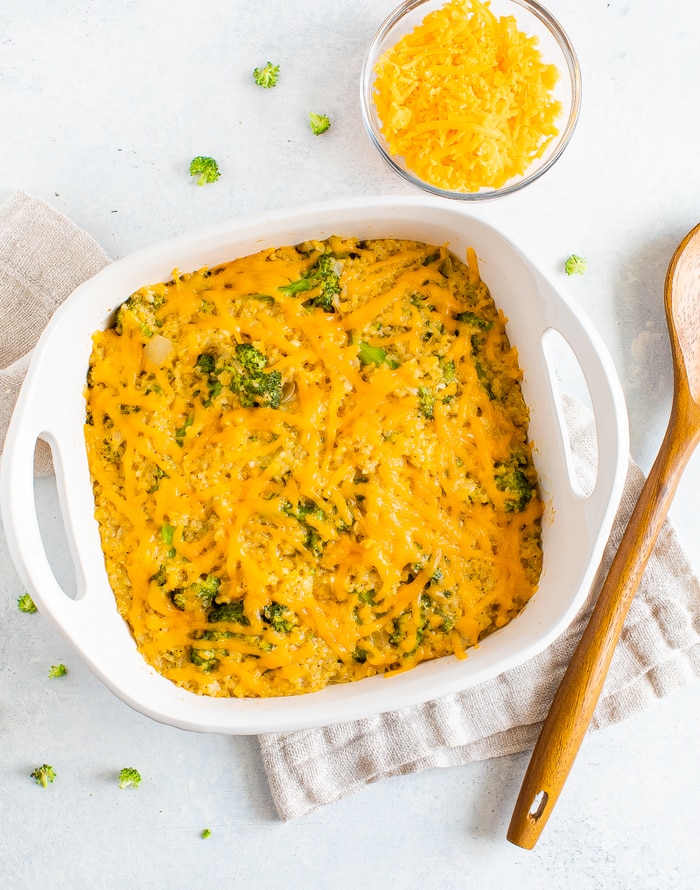 Baking dish with cheesy broccoli quinoa casserole. A wood spoon and dish towel are around the dish.