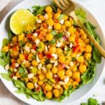 Bowl of curried chickpea salad made with chopped red onion, red pepper, cilantro, and dates, served over a bed of greens. Half a lime and gold fork also in the bowl.