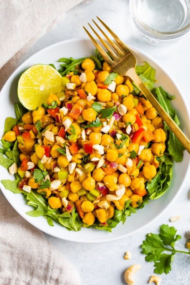 Bowl of curried chickpea salad made with chopped red onion, red pepper, cilantro, and dates, served over a bed of greens. Half a lime and gold fork also in the bowl.