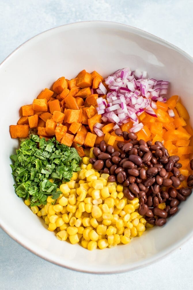 Ingredients for a southwestern quinoa salad in a bowl before being mixed: sweet potato, onion, peppers, black beans, corn, and cilantro.