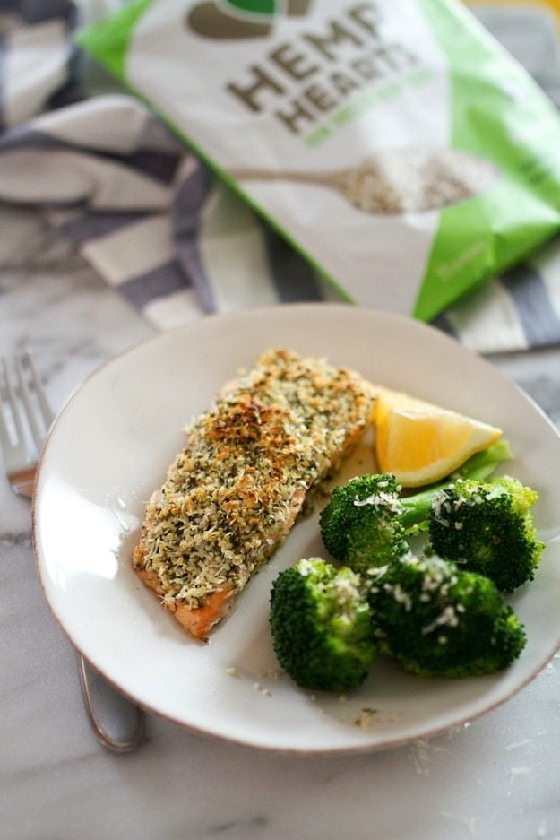 A white plate with a filet of hemp and pecorino crusted salmon served with a side of broccoli and a lemon wedge. A bag of Hemp Hearts is out of focus in the background.