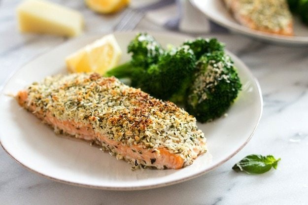 A close up of hemp and pecorino crusted salmon on a plate. Broccoli and a lemon wedge are out of focus on the plate.