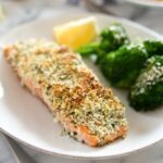 A filet of hemp and pecorino crusted salmon on a plate with broccoli and a lemon wedge out of focus on the plate.