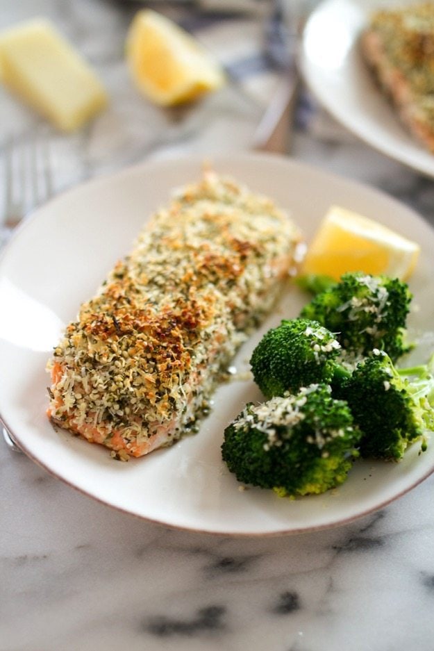 A white plate with a filet of hemp and pecorino crusted salmon served with a side of broccoli and a lemon wedge.