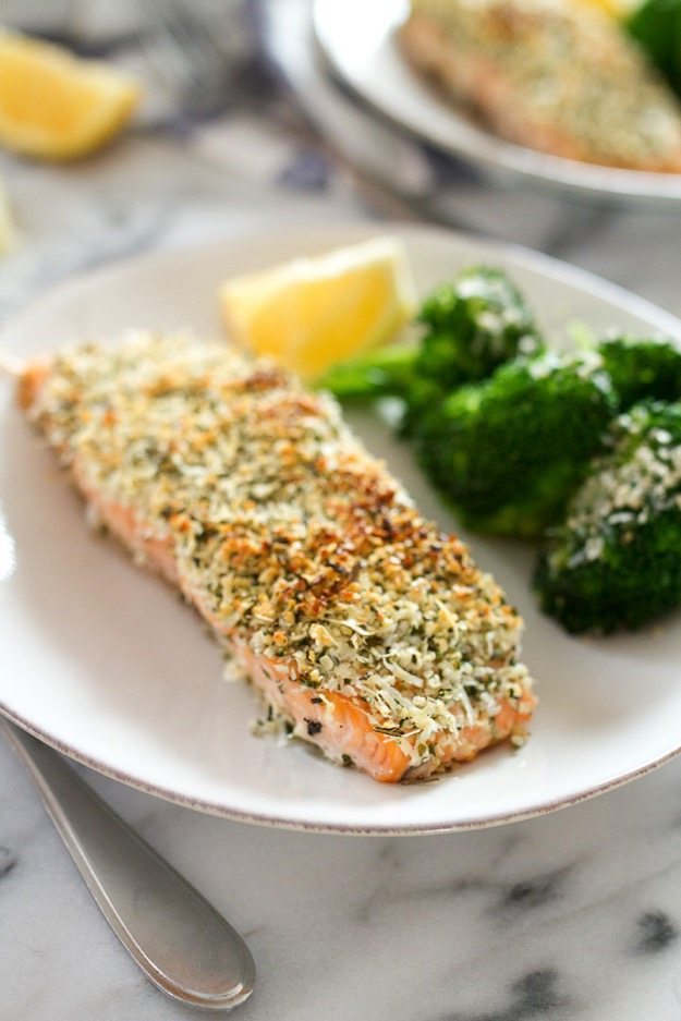 A filet of hemp and pecorino crusted salmon on a plate with broccoli and a lemon wedge out of focus on the plate.