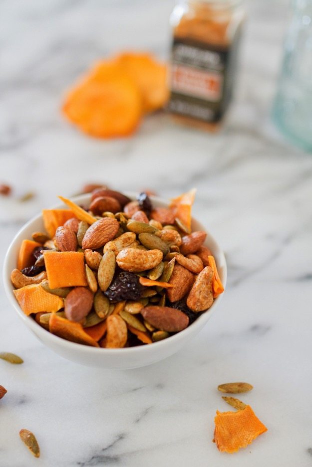 Mango curry trail mix in a white bowl on a marble counter with curry powder in the background. 