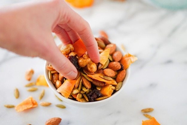 Mango curry trail mix in a white bowl on a marble counter with hand reaching in to grab some trail mix. 
